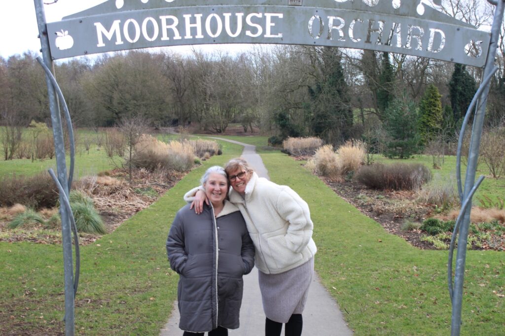 The two women stand under a decorative metal archway that reads "Moorhouse Orchard." They smile as they pose on a paved path surrounded by trees and shrubs.