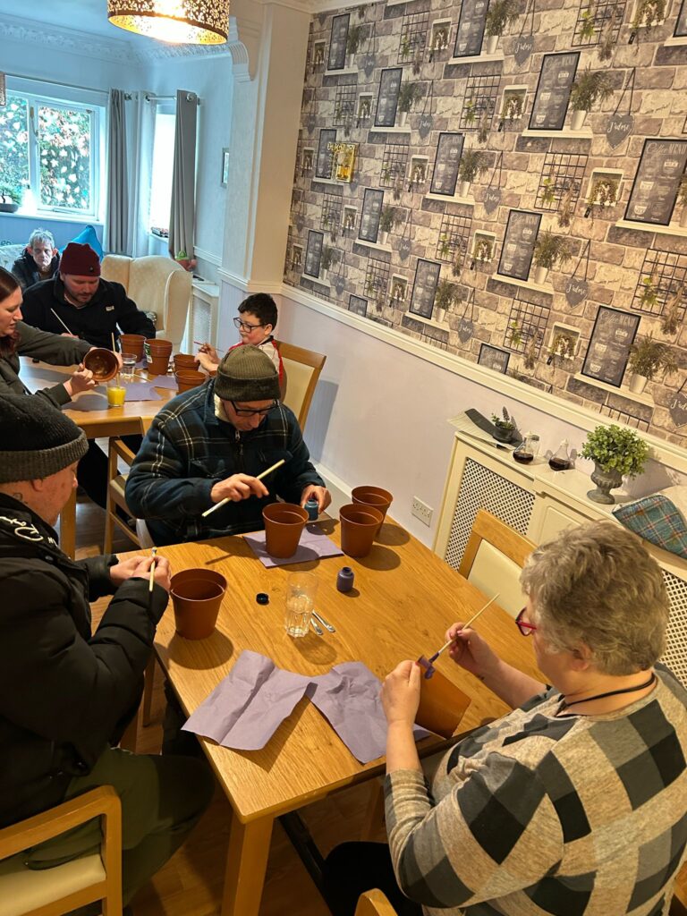 A wider shot of the room, showing multiple participants engaged in the pot painting activity. The group includes individuals of different ages, all focused on their creative work. The room has wooden flooring, a feature wall with decorative elements, and natural light coming in through the windows.