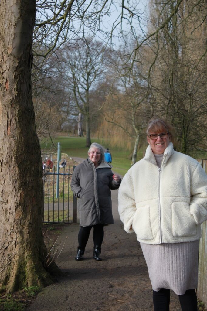 A candid moment of the two women walking in a park. One is closer to the camera in a cream fleece jacket, while the other follows behind in a grey coat. A tree trunk is in the foreground, with a park gate and pond in the background.