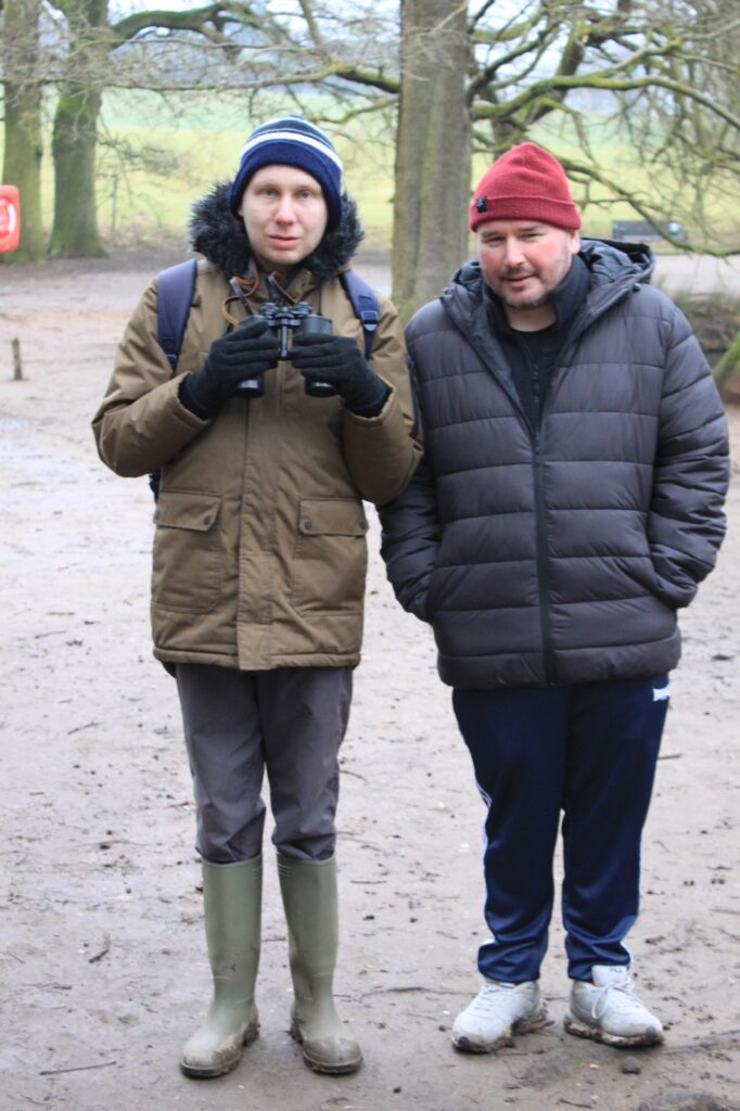 Two men posing for a photo, one holding binoculars and wearing wellies, while the other is dressed in a thick coat and beanie.