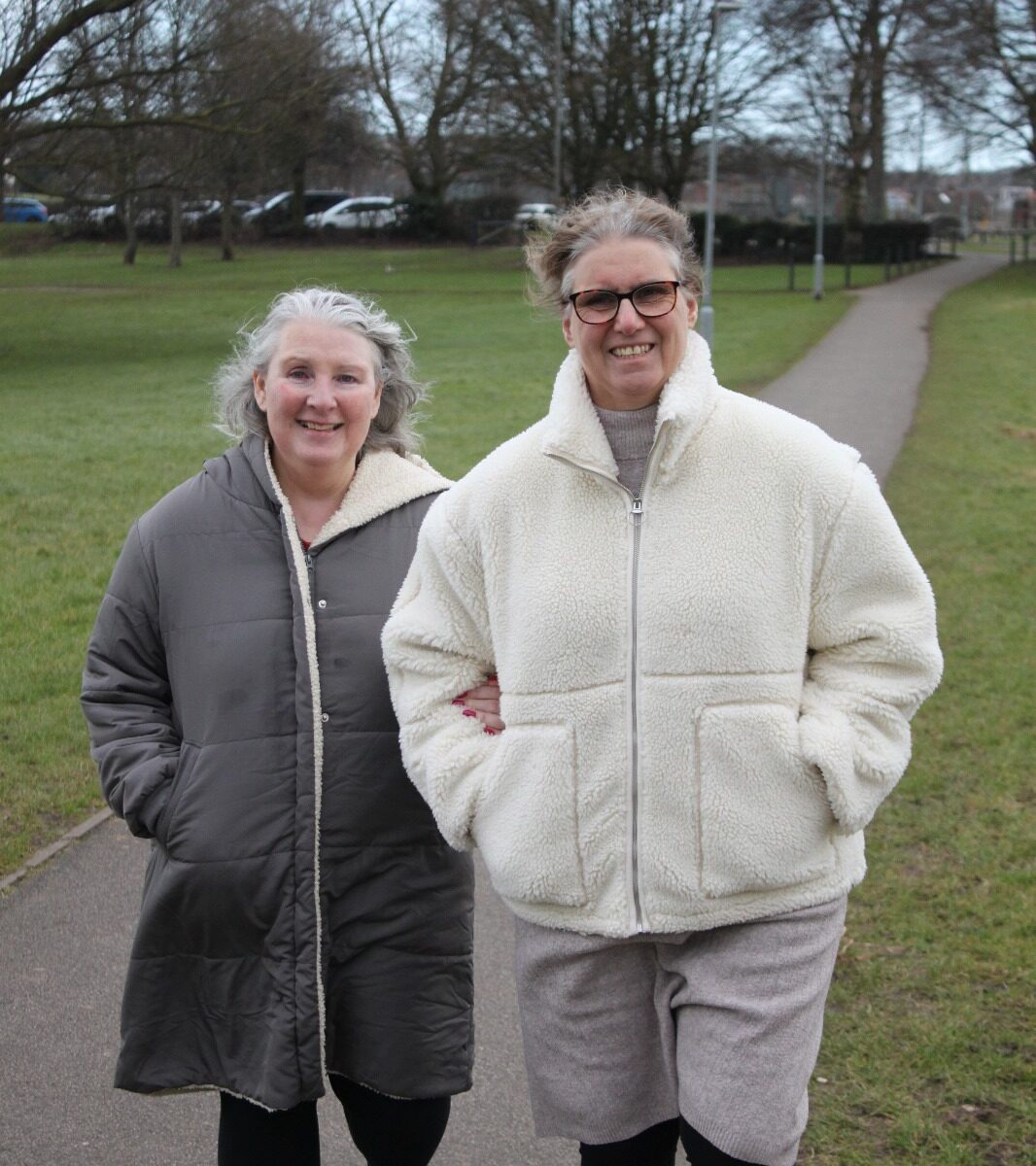 The same two women are walking arm in arm through an open park. They smile while strolling along a wide pathway lined with grass, trees, and distant houses.