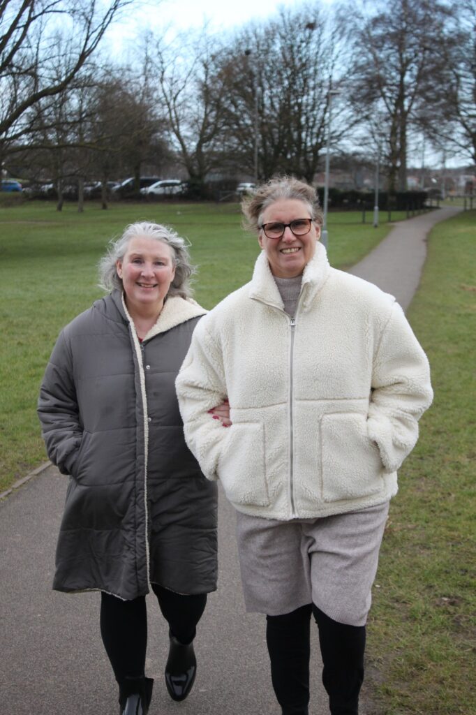 The same two women are walking arm in arm through an open park. They smile while strolling along a wide pathway lined with grass, trees, and distant houses.