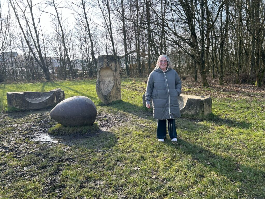 A smiling woman in a grey coat stands in a grassy area surrounded by wooden and stone sculptures, including a large carved seed and a tree trunk with an embedded relief carving.