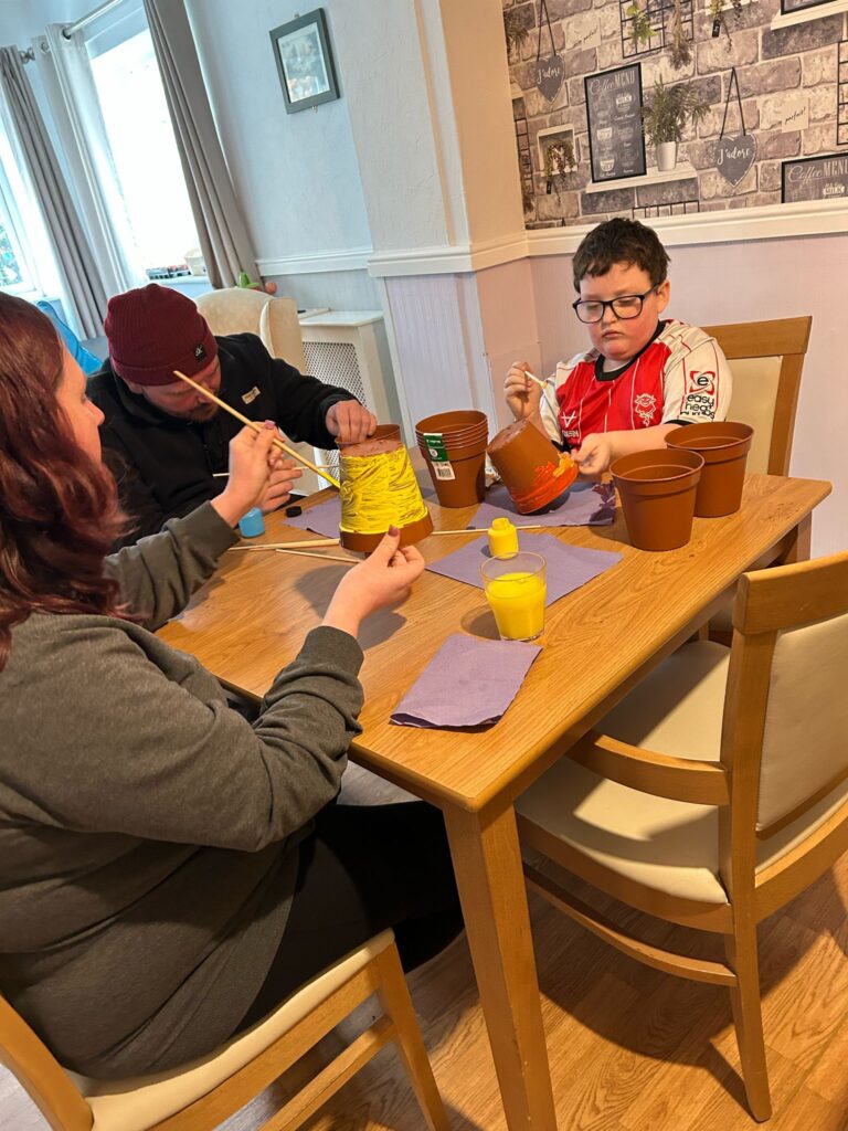 Another angle of the young boy painting his pot, with an adult next to him adding details to theirs. The warm-toned room has a patterned feature wall with decorative elements such as plants and framed prints, adding to the cosy atmosphere.