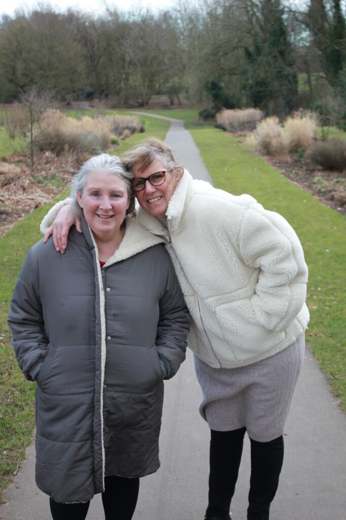 Two women stand together on a paved path in a park, smiling at the camera. One wears a grey coat, while the other wears a cream-coloured fleece jacket and glasses. They embrace warmly, with a backdrop of trees and grass.