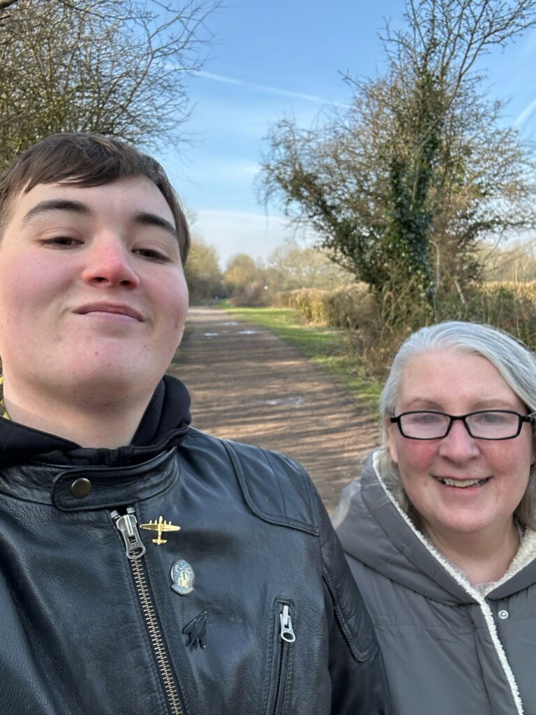 A close-up selfie of two people smiling outdoors, with a well-trodden walking path and trees in the background.