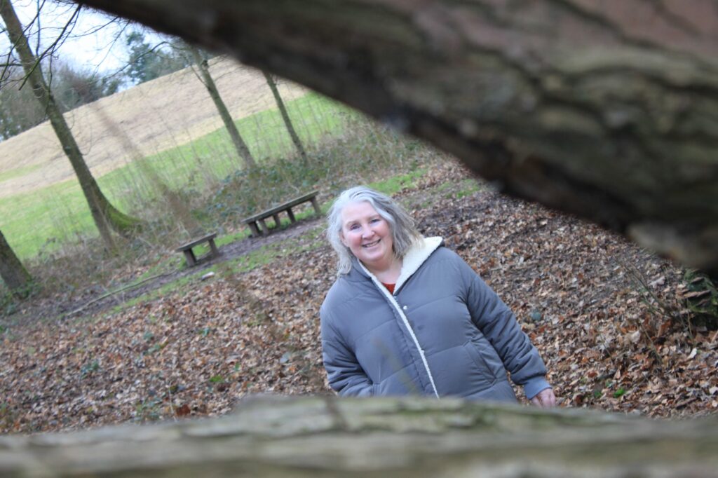 A woman in a grey coat is framed by the branches of a fallen tree, smiling as she stands in a wooded area covered in autumn leaves. Wooden benches are visible in the background.