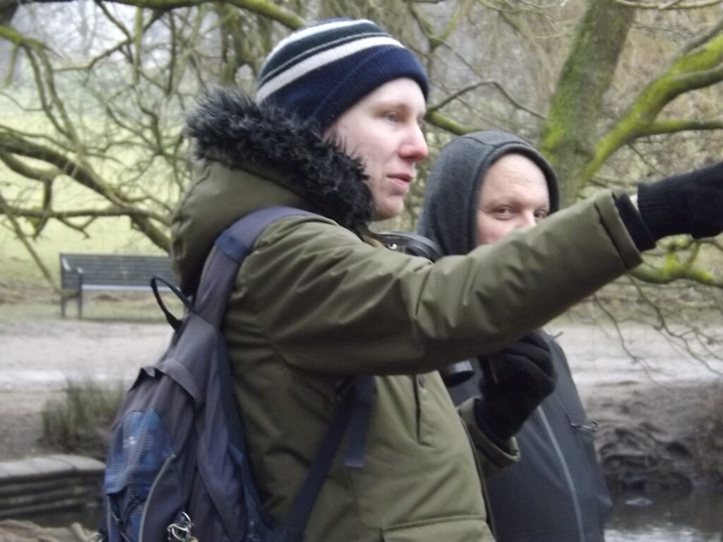 Two people standing near a tree, one pointing into the distance, both dressed warmly with backpacks.