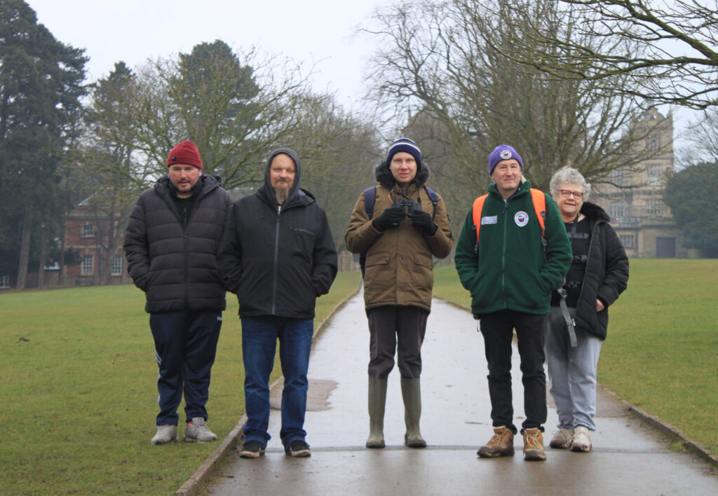 A group of five people in warm clothing standing on a path in Wollaton Park, with Wollaton Hall partially visible in the background.