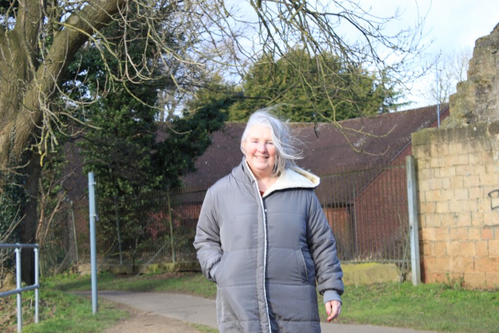 A woman with light skin and silver hair, wearing a grey coat, smiles while walking outdoors in a park. The wind blows her hair, and she is surrounded by trees and an old stone wall.