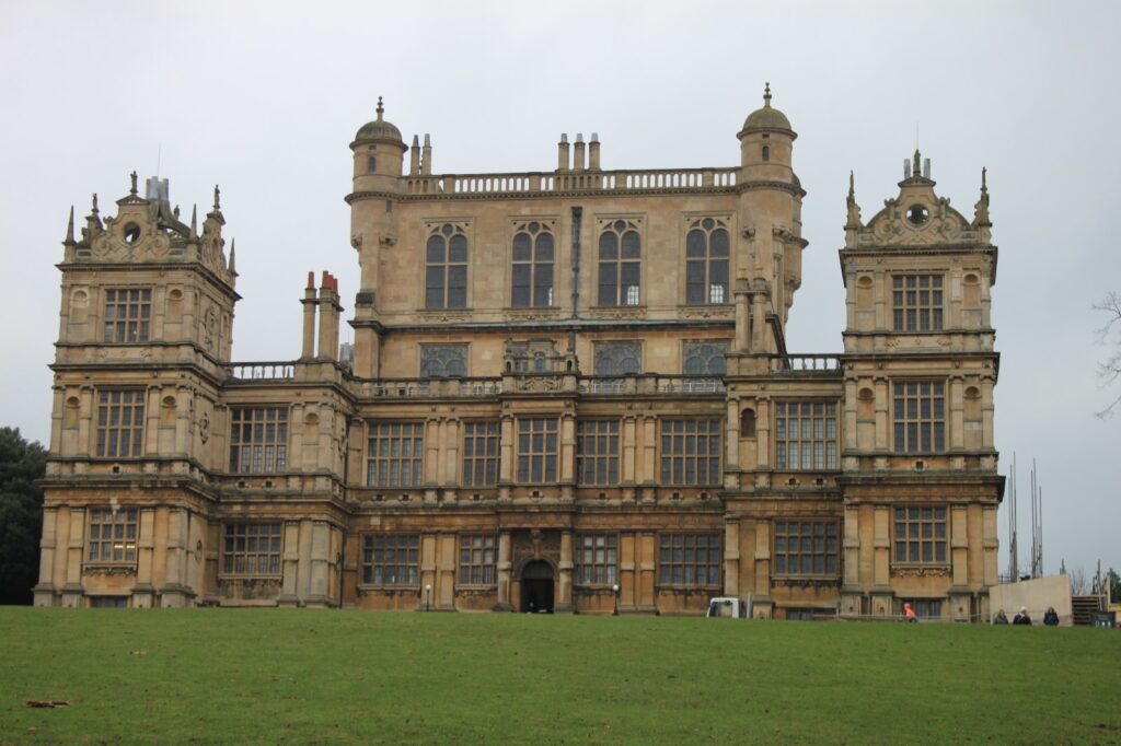 Wollaton Hall, a grand Elizabethan mansion with intricate architectural details, stands against an overcast sky with a large green lawn in the foreground.