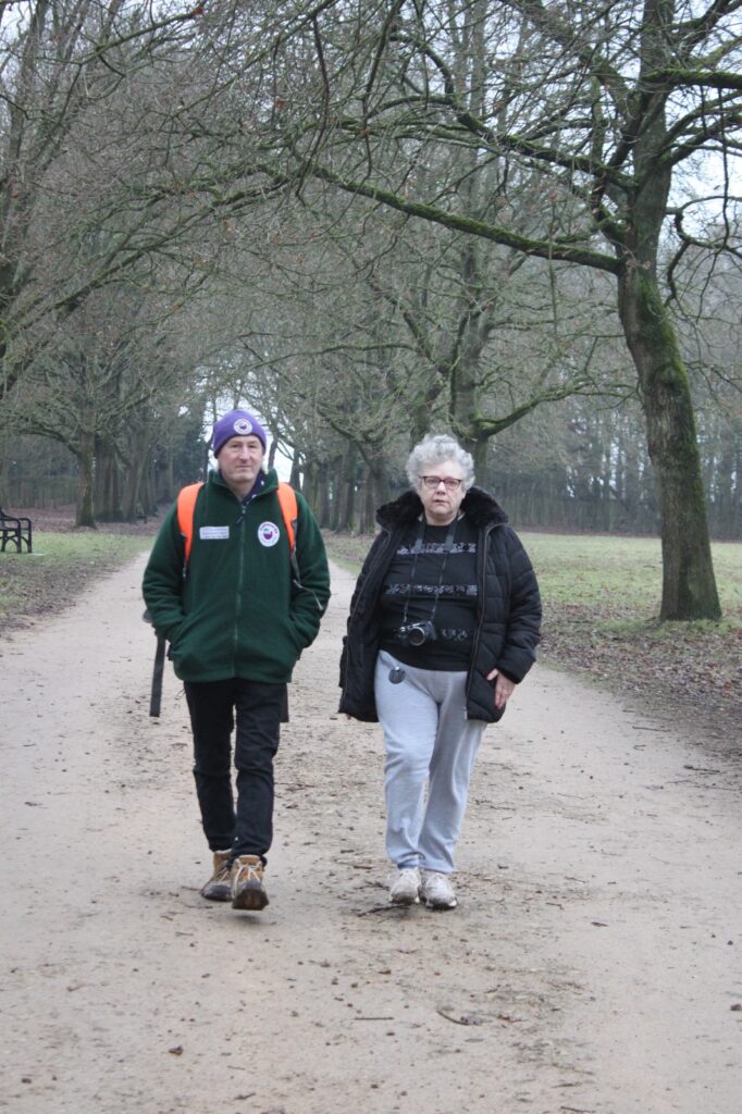 Two people walking along a tree-lined path in Wollaton Park, both wearing warm clothing. One is carrying a camera around their neck, and the other has a bright orange backpack.